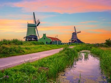 Traditionelle niederländische Windmühlen entlang eines Weges neben einem Kanal unter einem farbenfrohen Sonnenuntergangshimmel in den Niederlanden.