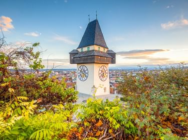 The Graz Clock Tower with white walls and dark pointed roof, framed by autumn foliage, overlooking the city at sunset.