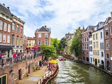 Gracht in Utrecht mit bunten historischen Häusern entlang des Wassers. Menschen sitzen auf einer Terrasse am Wasser unter gelben Sonnenschirmen.