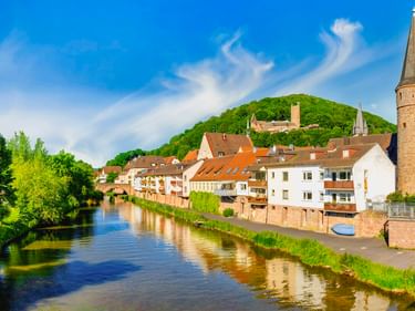 Historische Stadt Gemünden mit Fluss, traditionellen Häusern, Hexenturm mit Spitze und grünem Scherenburg-Hügel unter blauem Himmel.