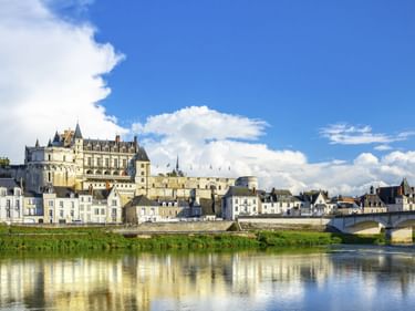 Schloss Amboise erhebt sich über der Stadt Amboise mit weißen Gebäuden entlang der Loire. Eine Steinbrücke überquert das Wasser unter blauem Himmel.