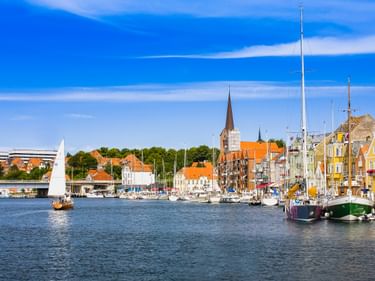 Sonderborg Hafen mit Segelbooten am Kai. Bunte historische Gebäude und ein Kirchturm säumen den Hafen unter blauem Himmel.