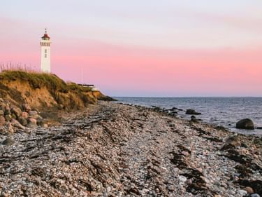 Weißer Leuchtturm Helnæs auf einer grasbewachsenen Klippe über einem Steinstrand bei Sonnenuntergang. Rosa Himmel über dem Meer auf Fünen.