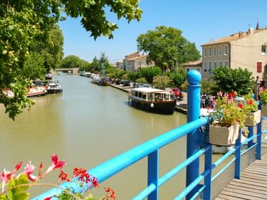Kanal bei Homps mit vertäuten Booten und blauem Geländer, das mit Blumen geschmückt ist. Traditionelle französische Gebäude säumen den Wasserweg.