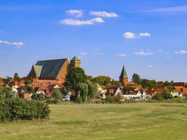 View of Verden with large church and tower rising above traditional red-roofed houses. Green meadow in foreground under blue sky.