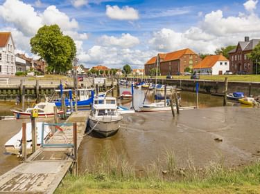Hafen von Tönning bei Ebbe mit kleinen Booten an Holzstegen. Traditionelle Häuser mit orangefarbenen Dächern säumen das Ufer unter blauem Himmel.