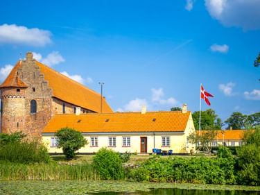 Nyborg Slot in Dänemark mit orangefarbenen Ziegeldächern, gelben Gebäuden und dänischer Flagge am Wasser unter blauem Himmel.