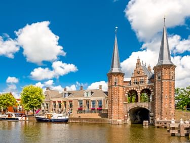 Das Wassertor in Sneek mit zwei Backsteintürmen und spitzen Turmhelmen über einem Kanal. Historische Gebäude und Boote säumen das Ufer unter blauem Himmel.