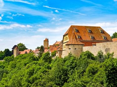 Mittelalterliche Gebäude der Altstadt Rothenburg mit orangefarbenen Ziegeldächern, Steinmauern und Türmen über dichtem grünem Wald.