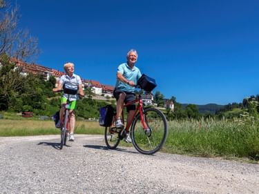 Zwei Radfahrer auf einem Schotterweg mit grünen Wiesen und einem Dorf mit roten Dächern am Hang unter blauem Himmel.