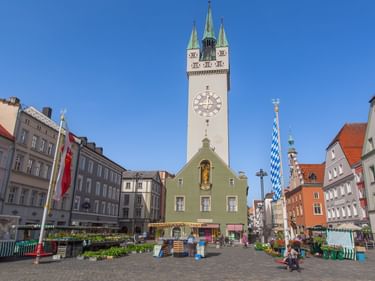 Straubing town square with tall clock tower, green building, Bavarian flag, and colorful historic buildings under blue sky.