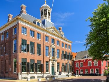 Rotes Backsteinrathaus in Leeuwarden mit verziertem Glockenturm, weißen Fensterläden und dekorativem Eingang. Angrenzende rote Gebäude und grüner Baum.