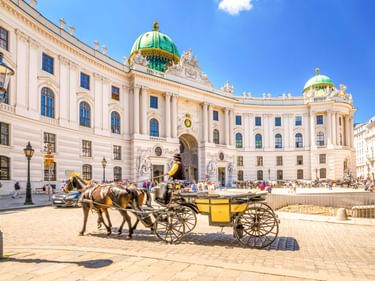 Gelber Fiaker vor der Alten Hofburg in Wien mit ihrer weißen Barockfassade und den charakteristischen grünen Kuppeln.