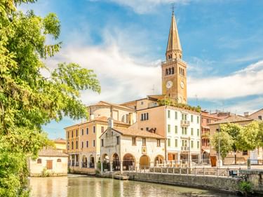 Der Glockenturm des St. Andreas Doms erhebt sich über bunte Gebäude entlang eines Kanals in Portogruaro, eingerahmt von grünen Bäumen.