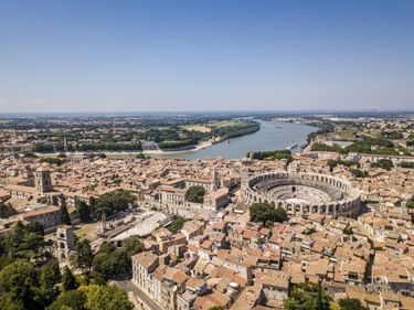 Luftaufnahme von Arles, Frankreich, mit dem historischen römischen Amphitheater, umgeben von Gebäuden mit Terrakottadächern und der Rhône im Hintergrund.