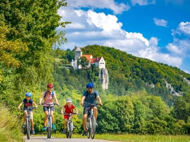 Vierköpfige Familie radelt auf einem Weg mit Burg Prunn auf einem bewaldeten Hügel. Blauer Himmel mit weißen Wolken über grüner Landschaft.