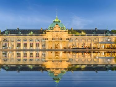 Kaiserpalais in Bad Oeynhausen, a grand yellow palace with green dome reflected in a pond. The neoclassical building is framed by trees.
