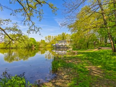 Grüner Uferweg an der Elbe in Torgau mit Bäumen und einem Raddampfer, der sich im ruhigen Wasser unter blauem Himmel spiegelt.