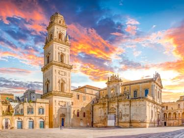 Kathedrale von Lecce mit hohem Glockenturm im Barockstil unter dramatischem orange-blauem Sonnenuntergangshimmel. Der historische Platz ist gepflastert.