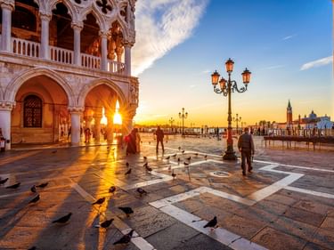 Markusplatz bei Sonnenaufgang mit Dogenpalast, verzierten Laternen, Tauben auf gemustertem Pflaster und gehenden Menschen. Goldenes Licht beleuchtet die Szene.