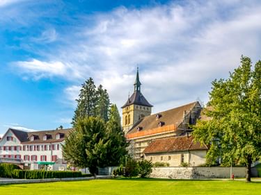 Kirche mit spitzem Turm und historische Gebäude in Arbon, umgeben von grüner Wiese und Bäumen unter blauem Himmel mit weißen Wolken.