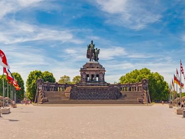 Deutsches Eck Denkmal in Koblenz mit Reiterstatue auf Steinsockel, umgeben von bunten Flaggen und Flussblick unter blauem Himmel.