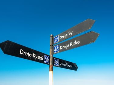 Radweg-Wegweiser auf der Insel Drejø mit Richtungen nach Drejø By, Drejø Kirke und Drejø Kystli vor klarem blauen Himmel.