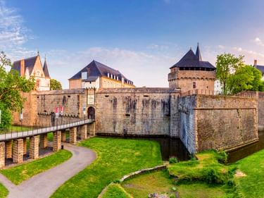 Schloss der Herzöge der Bretagne in Nantes mit Steinmauern, Türmen und Wassergraben. Eine Brücke führt zum Eingang, umgeben von grünen Rasenflächen.