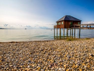 Hölzernes Badehaus auf Stelzen am Bodensee in Bregenz. Kieselstrand im Vordergrund, ruhiges Wasser und blauer Himmel mit Wolken im Hintergrund.