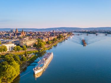 Aerial view of Mainz with the Rhine River flowing through. Cruise ships docked along the riverbank, historic city center with church spires visible.