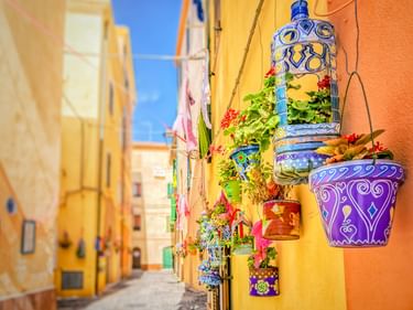 Enge Gasse in Alghero mit gelben und orangefarbenen Gebäuden, geschmückt mit bunten Keramiktöpfen und Blumen an den Wänden.