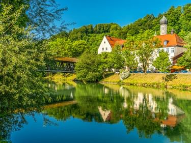 St. Andreas Kirche mit weißem Turm und rotem Dach neben einem ruhigen Fluss in Wolfratshausen, Bayern. Eine Holzbrücke überquert das Wasser, umgeben von üppigem Grün.