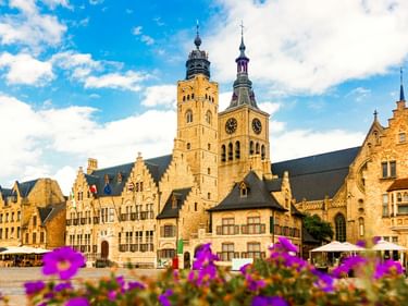 Historischer Grote Markt in Diksmuide mit flämischen Giebelhäusern, Kirchtürmen und lila Blumen im Vordergrund unter blauem Himmel.