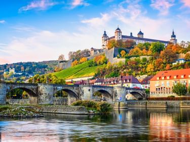 Festung Marienberg auf einem Hügel mit herbstlichen Weinbergen, alte Steinbrücke über den Main und bunte Gebäude in Würzburg bei Sonnenuntergang.