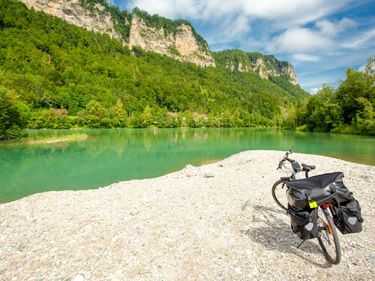 Tourenrad mit Packtaschen auf Kiesbank am türkisfarbenen Drauufer in Österreich, mit bewaldeten Klippen im Hintergrund.