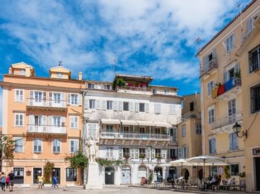 Sonniger Platz in Korfu-Stadt mit bunten mediterranen Gebäuden, Café-Außenbestuhlung und Fußgängern unter blauem Himmel.