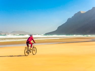 Radfahrer in pinker Kleidung fährt auf goldenem Sand am Strand Playa de Famara mit Meereswellen und dramatischen Klippen im Hintergrund.