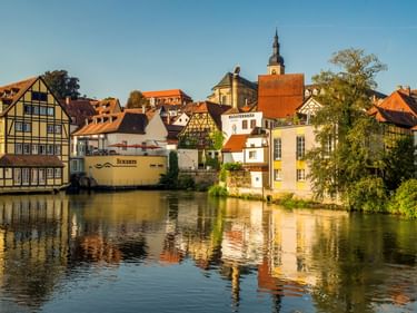 Colorful half-timbered houses and historic buildings of Bamberg's old town reflected in the river, with a church tower visible in the background.