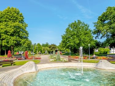 Runder Brunnen mit Wasserfontänen im Bad Füssing Kurpark, umgeben von bunten Blumenbeeten, Bänken und hohen Bäumen unter blauem Himmel.