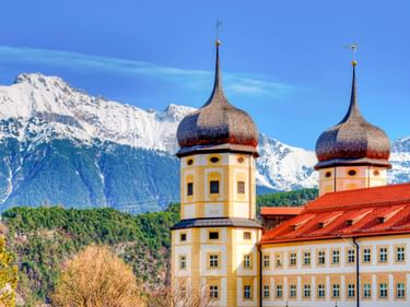 Stift Stams mit zwei Zwiebeltürmen und gelber Fassade vor schneebedeckten Alpengipfeln in Tirol, Österreich.
