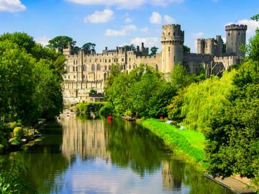 Warwick Castle mit Steintürmen und Mauern an einem ruhigen Fluss, umgeben von üppigen grünen Bäumen und Rasenflächen unter blauem Himmel.
