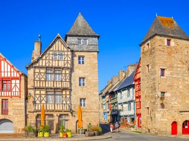 Mittelalterliche Fachwerkhäuser und Steintürme in der Altstadt von Tréguier unter blauem Himmel. Bunte Fassaden mit rot-weißen Mustern säumen den Platz.