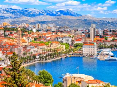 Panoramablick auf den Hafen von Split in Kroatien mit rotgedeckten Gebäuden, palmengesäumter Uferpromenade, Booten im blauen Wasser und schneebedeckten Bergen.