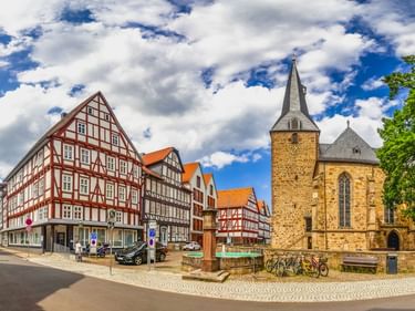 Steinkirche mit spitzem Turm neben bunten Fachwerkhäusern auf dem Marktplatz der Altstadt Melsungen unter blauem Wolkenhimmel.