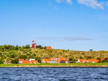 Rot-weißer Leuchtturm auf einer Düne mit Blick auf Häuser mit orangen Dächern auf der Insel Vlieland. Blaues Wasser im Vordergrund.