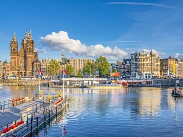 Amsterdamer Grachtenblick mit den Zwillingstürmen der Nikolausbasilika, historischen Gebäuden und Bootsanlegern unter blauem Himmel.
