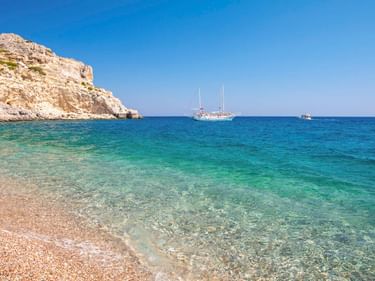 Afandou Strand mit kristallklarem türkisfarbenem Wasser, Kieselstrand, Felsenklippen links und einem weißen Segelboot im ruhigen Meer.