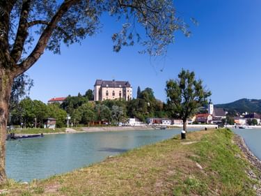 View of Grein Castle on a hill above the Danube River. Green riverbank with trees in foreground, white church tower and hills in background.
