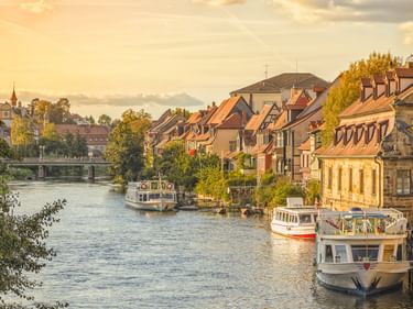 Reihe bunter historischer Häuser entlang eines Flusses in Bayern mit vertäuten Booten. Eine Brücke und Kirchturm sind im Hintergrund im goldenen Licht sichtbar.