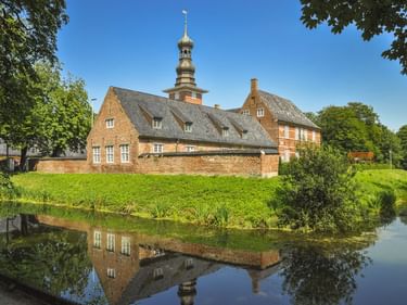 Schloss Husum, ein rotes Backsteingebäude mit markanten Turm, umgeben von grüner Wiese und Wassergraben. Das Gebäude spiegelt sich im Wasser.